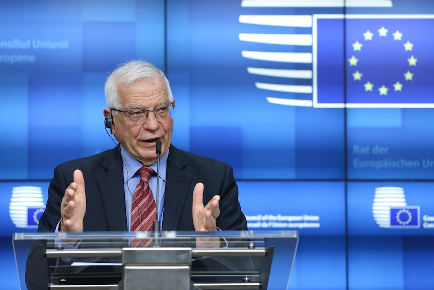 A grey-haired man presents at a lectern in front of an EU banner
